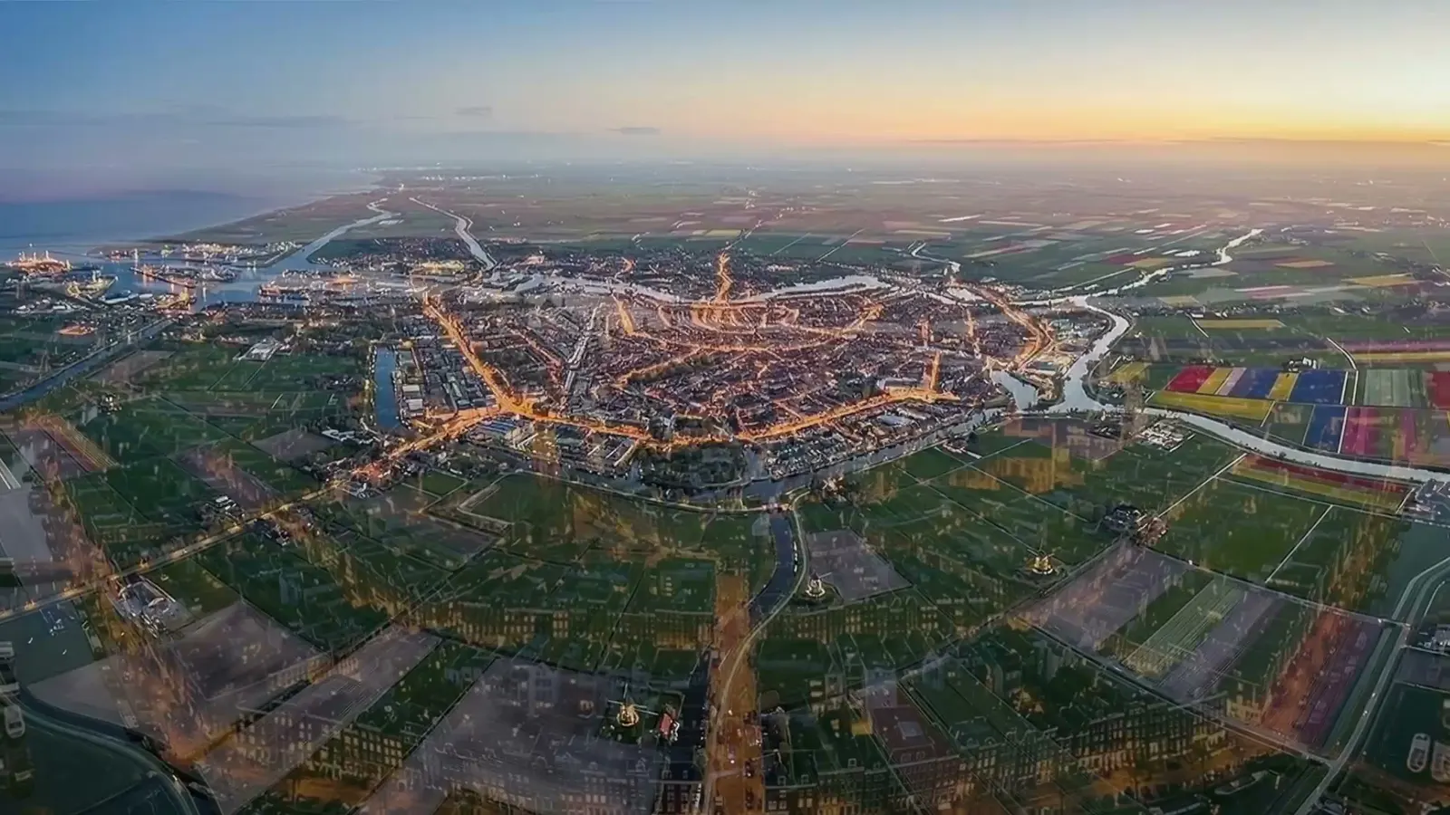 Amsterdam from high above at dusk, the canal ring lit warm orange against blue water, river and coast at the top of frame.