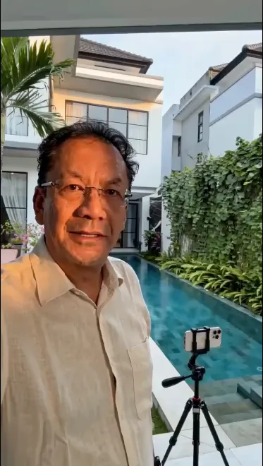 The presenter standing in front of a villa and its pool in soft daylight, a phone on a tripod set up facing them.