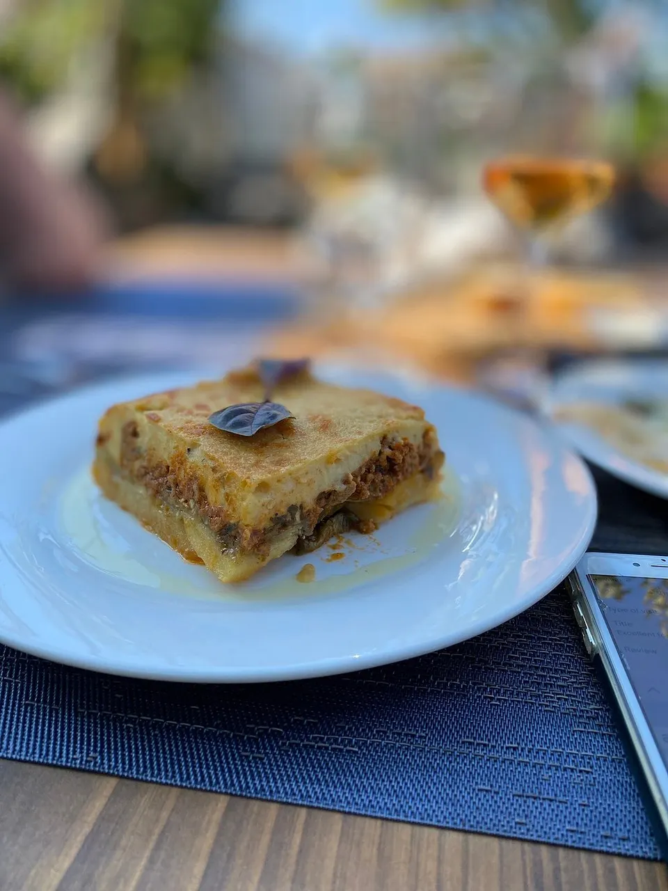 A slice of moussaka on a white plate, photographed on the restaurant terrace in daylight with a wine glass behind it.