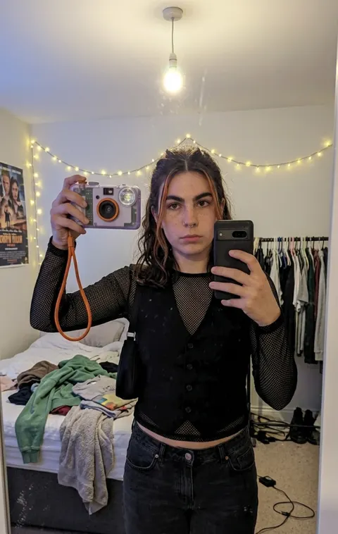 A woman taking a mirror selfie in a bedroom strung with fairy lights, holding a small compact camera on an orange wrist strap.