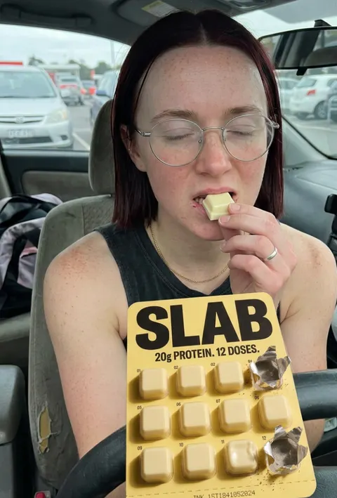 A woman in a car park eating a SLAB block, holding the yellow tray up to camera with the pack claim facing forward.
